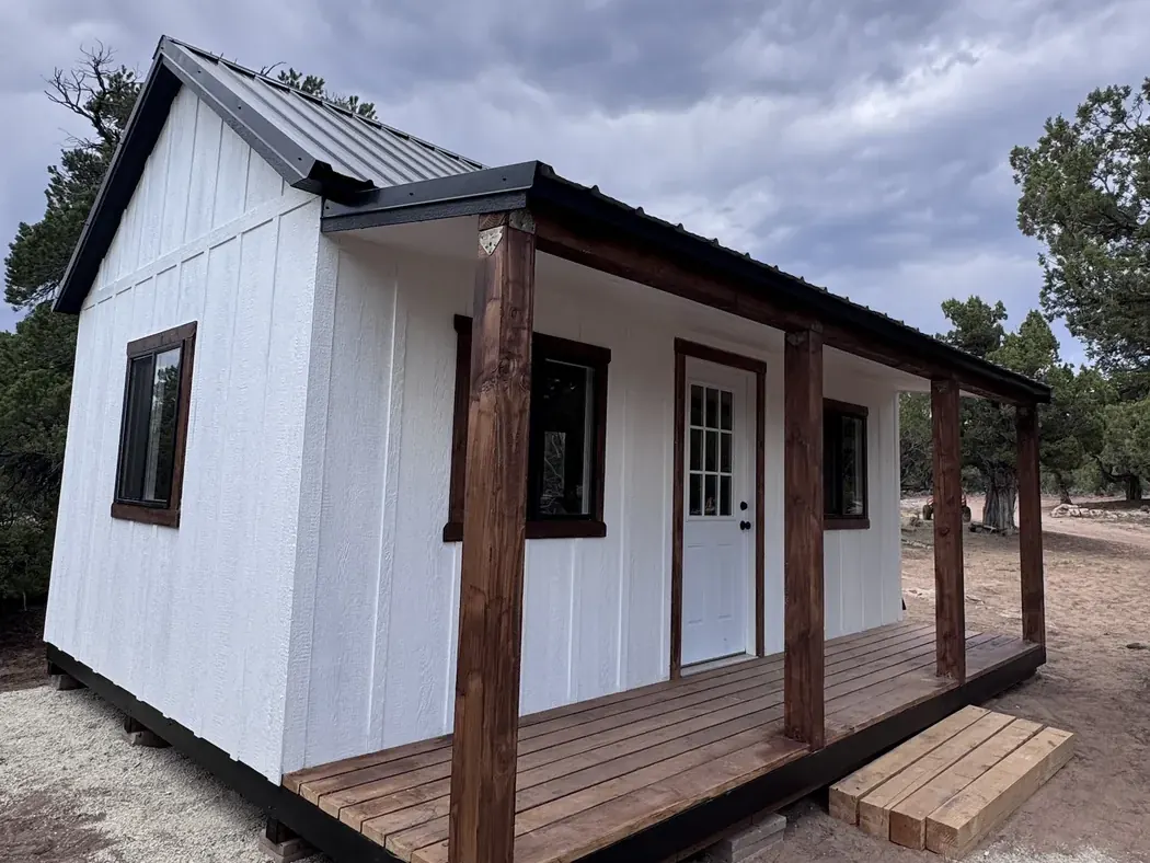 White cabin style shed with a full cedar front porch and a dark metal roof, built by ShedPro LLC
