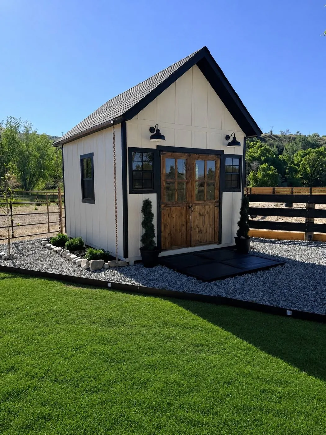 White board-and-batten cottage shed with a steep gable roof, cedar french doors, black trim and barn lights, built on-site by ShedPro LLC